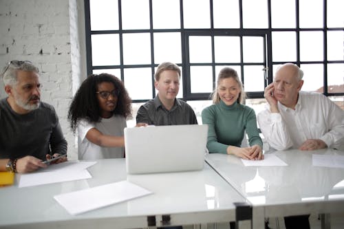 Group of multiracial coworkers of different ages discussing details of project while using computer at table in modern workplace