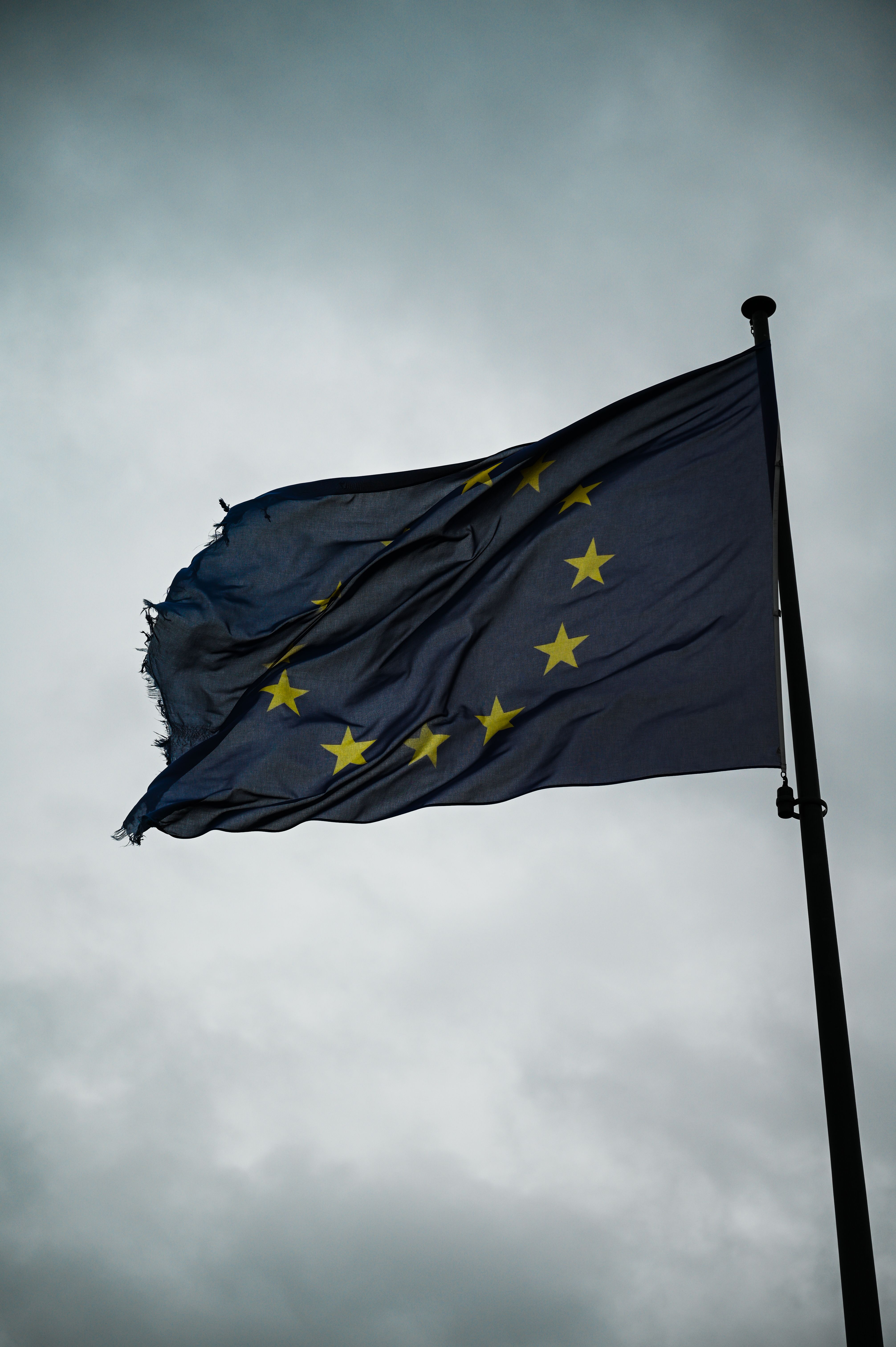 A tattered European Union flag waving against a moody, cloudy sky in Brussels.