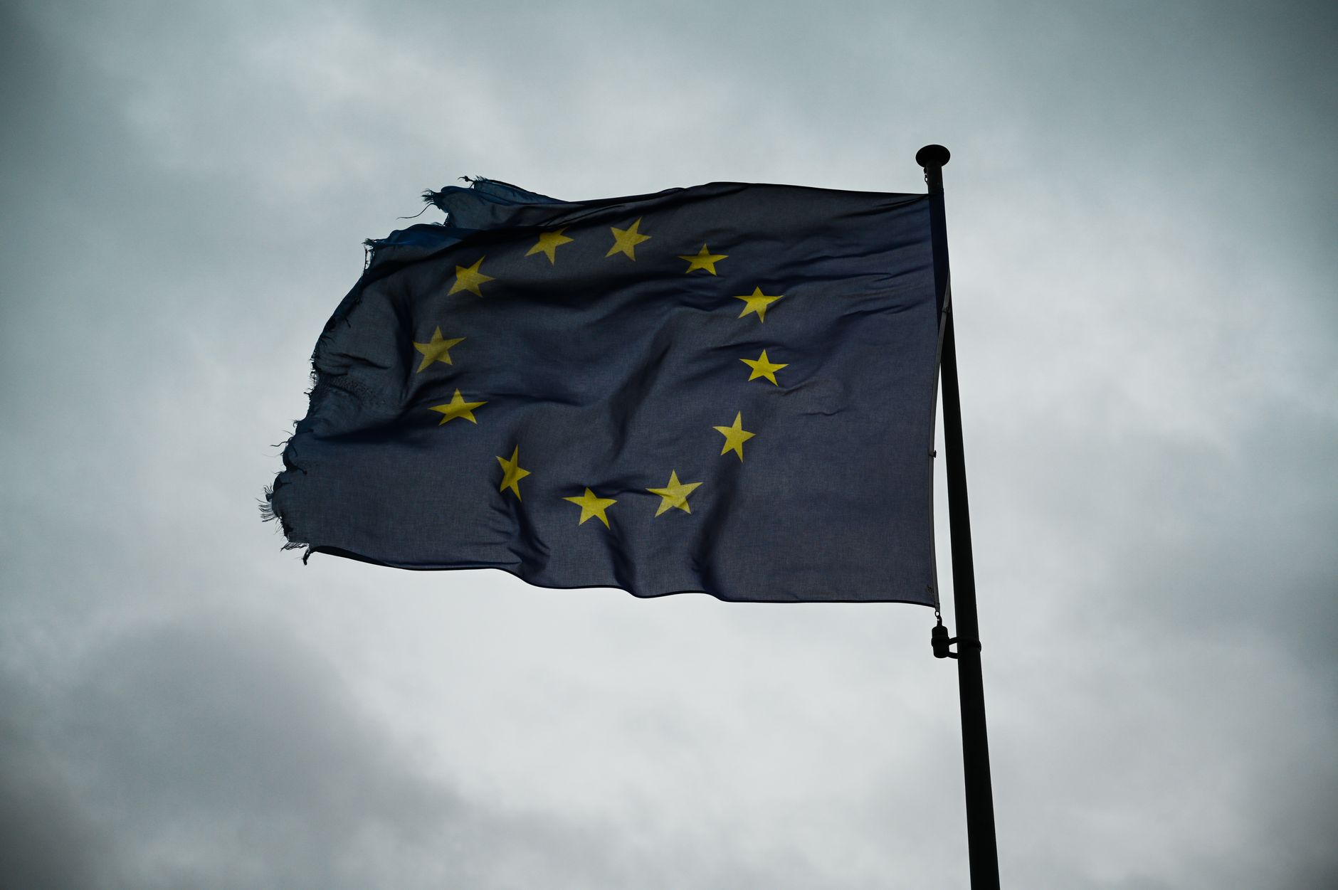 A frayed European Union flag waves on a cloudy day, symbolizing unity and resilience.