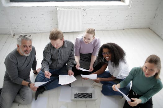 A diverse team of adults engaged in collaborative work, seated indoors with laptops and documents.