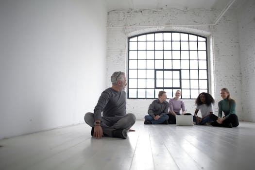 Full body of positive multiracial colleagues in casual clothes sitting in lotus pose on floor while resting together during break using laptop in room