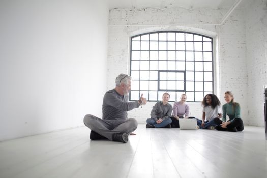 Full body of mature male employee holding thumb up during training with young diverse coworkers sitting on floor with crossed legs in room