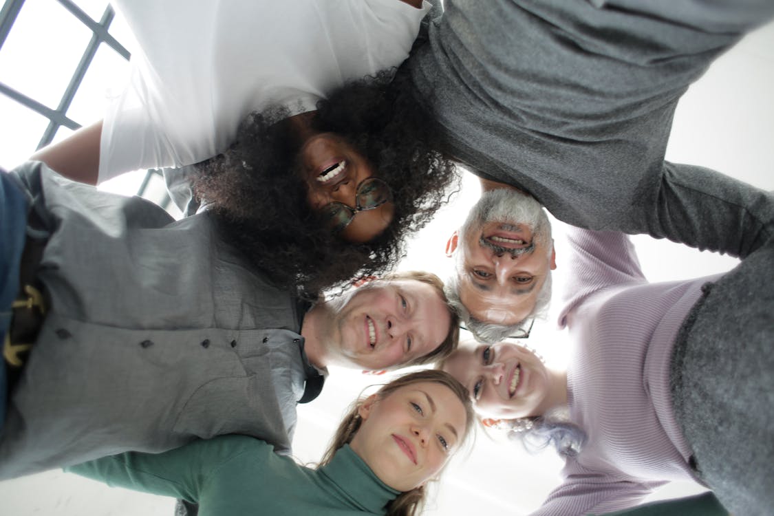 Free Multiethnic colleagues of different ages in casual wear standing in circle and hug each other smiling at camera in light workspace Stock Photo