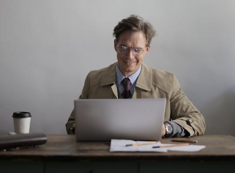 Confident businessman using laptop in office, enjoying a beverage and smiling.