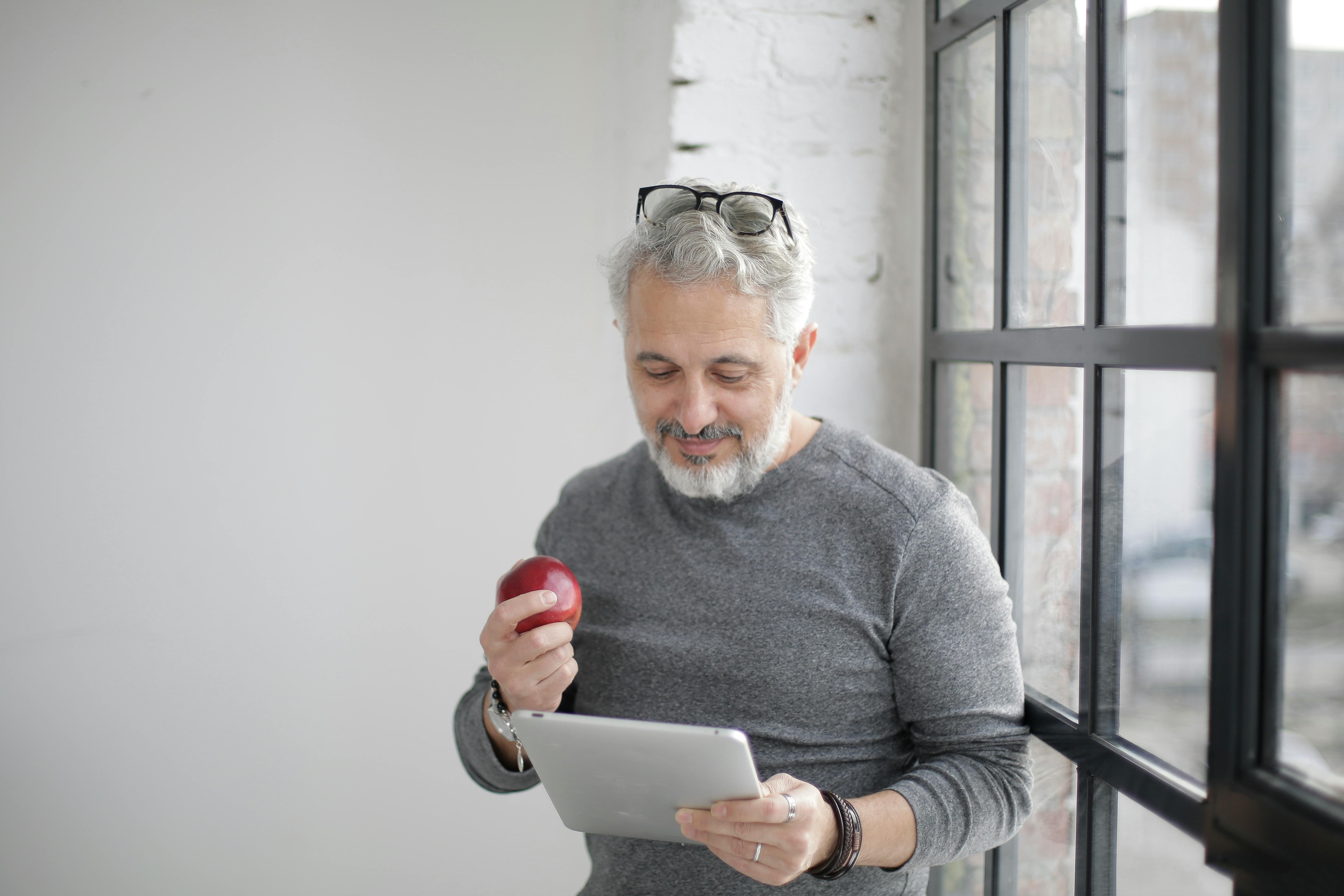 positive mature employee surfing tablet while eating apple in light workplace