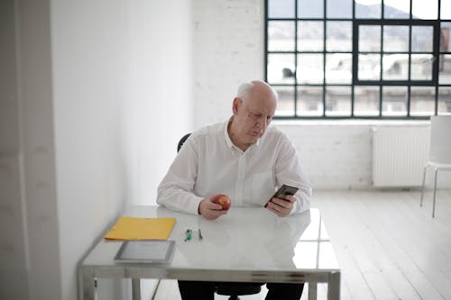 Free Elderly man in white shirt holding apple and messaging on mobile phone during short break in contemporary office Stock Photo