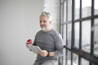 Smiling middle aged man using tablet and holding apple near window in office