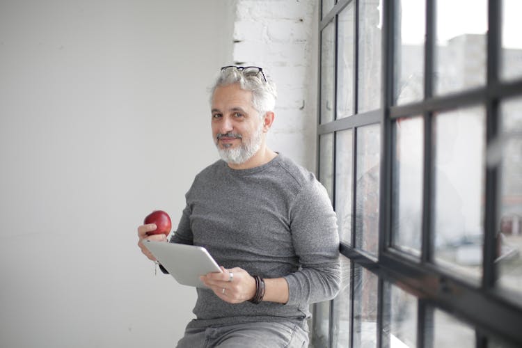 Smiling Middle Aged Man Using Tablet And Holding Apple Near Window In Office