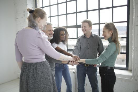 Happy multiethnic coworkers of different ages in casual wear smiling and joining hands together before working day in contemporary workplace against window