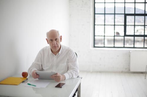 Senior male professor in white shirt using tablet while sitting at table with smartphone documents and apple aside in spacious office