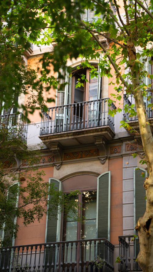 Free Beautiful façade of a Barcelona building with classic balconies, framed by lush green trees. Stock Photo