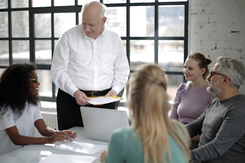 Positive multiracial colleagues of different ages gathering at table in modern workspace while old man presentation project