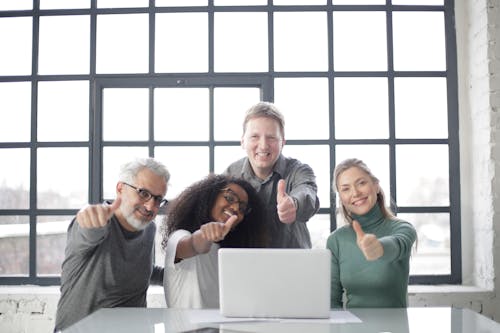 Free Happy multiracial team sitting at table with laptop and working on successful project while demonstrating thumb up gesture and smiling at camera Stock Photo
