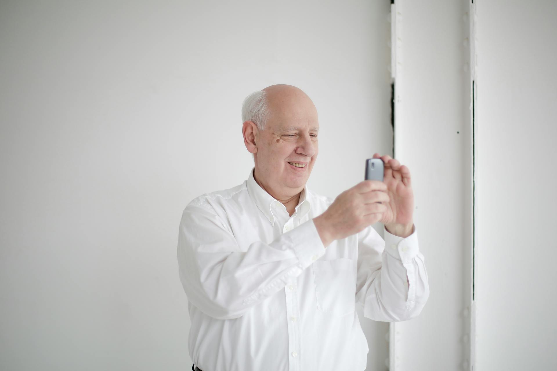 Positive old bald man in formal wear taking photo on cellphone while standing against white wall and toothy smiling