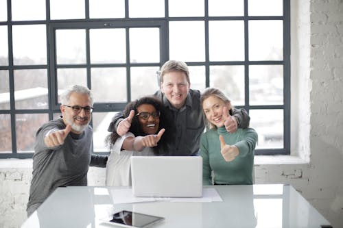 Cheerful diverse coworkers of different ages working on laptop on project while showing thumb up gesture and smiling when looking at camera