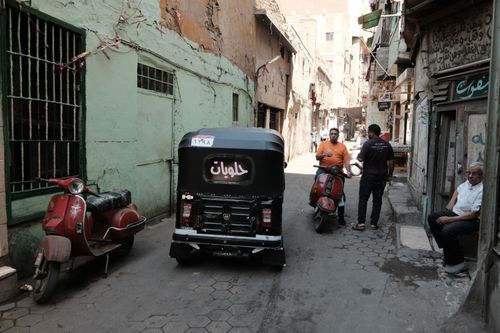 Free A bustling alleyway in Cairo featuring a tuk-tuk, scooters, and locals on a sunny day. Stock Photo