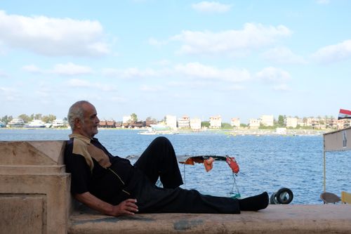 Free An elderly man enjoys a peaceful moment by the Nile River with a scenic view of Cairo's cityscape. Stock Photo