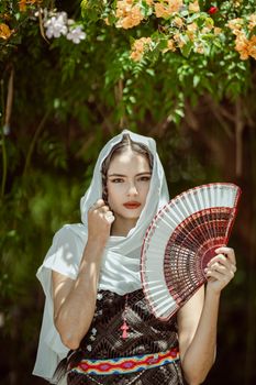 Woman in traditional attire holding a fan under vibrant flowers, shot in León, Mexico.