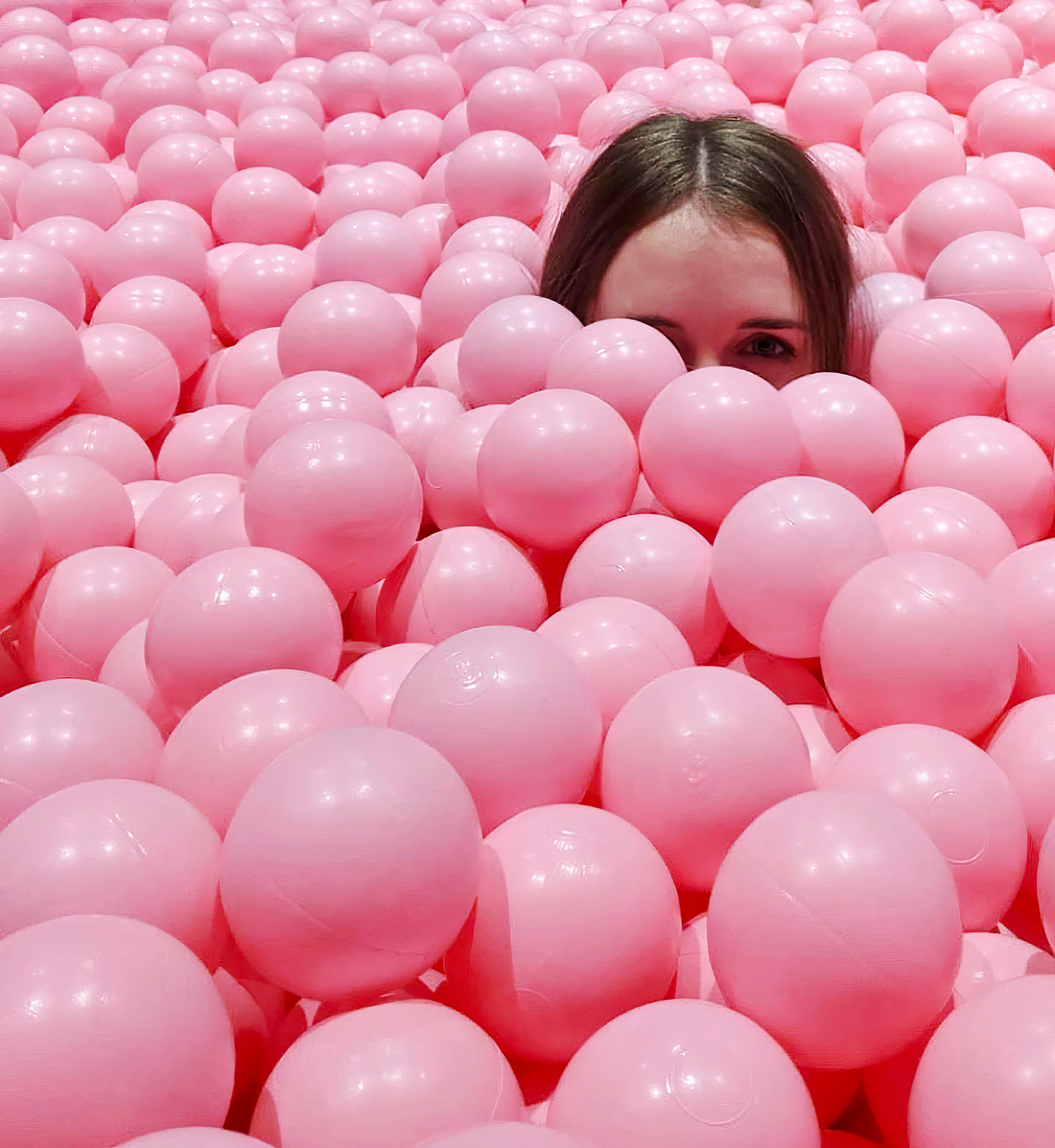 A young woman peeks through a sea of pink balls, creating a playful and lighthearted scene.