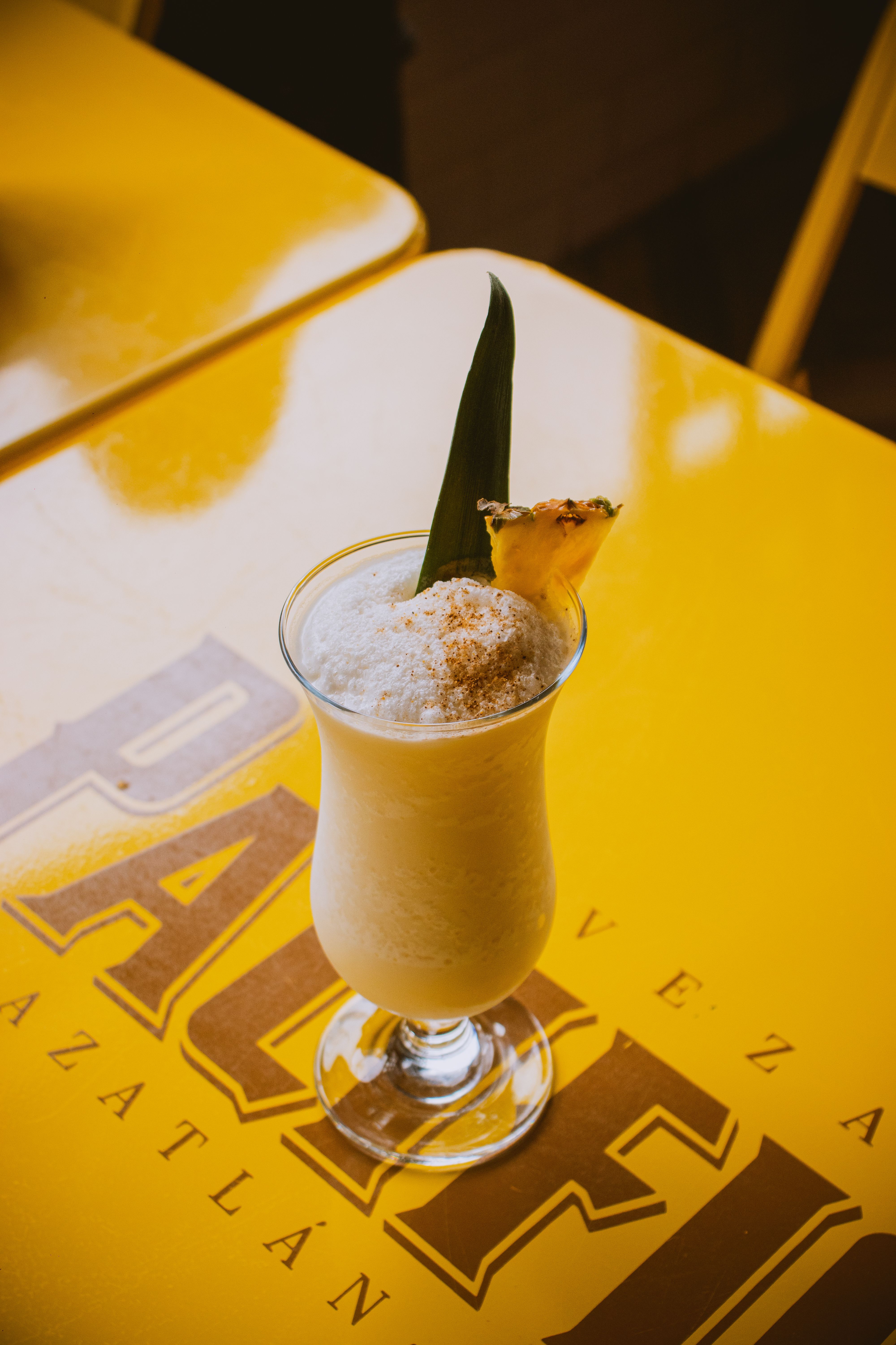 A tall glass of pale orange Painkiller cocktail over crushed ice, topped with a dusting of grated nutmeg and garnished with a pineapple wedge.