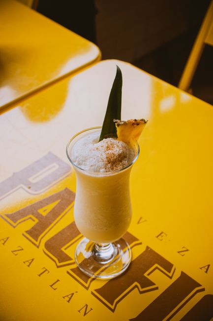 A tall glass of pale orange Painkiller cocktail over crushed ice, topped with a dusting of grated nutmeg and garnished with a pineapple wedge.