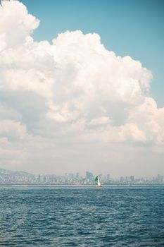 A sailboat in a calm sea under fluffy clouds with a city skyline in the distance.