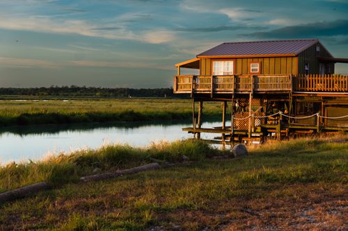 Free A tranquil riverside cabin surrounded by lush greenery during sunset in a serene countryside setting. Stock Photo