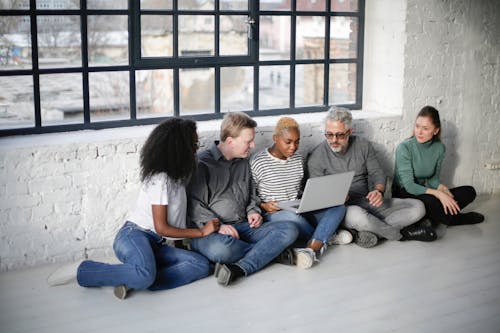 Focused group of diverse people sitting on floor with crossed legs while using laptop and working on project in loft styled workspace