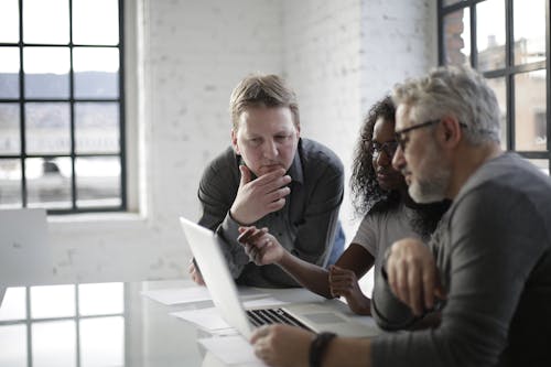 Free Concentrated serious multiracial people discussing details of creative project while sitting at table and using laptop in modern loft styled office Stock Photo