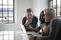 Concentrated serious multiracial people discussing details of creative project while sitting at table and using laptop in modern loft styled office
