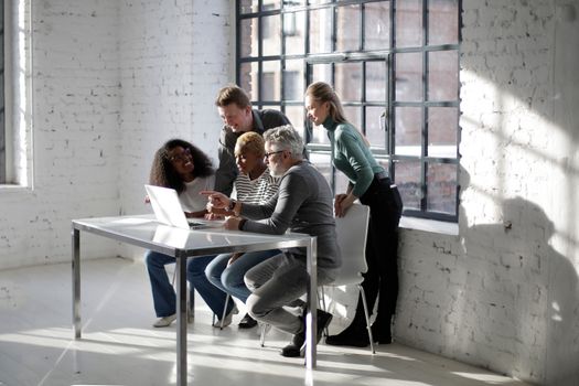 Group of multiracial coworkers of different age in casual outfit gathering at table with netbook and working on business project
