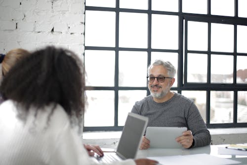 Cheerful aged gray haired man with tablet in casual clothes sitting with coworkers with laptop while working on project together