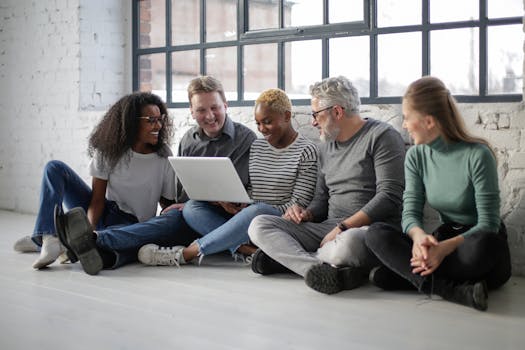 Happy creative multiethnic team with laptop sitting on floor in office with white brick walls and big window in daylight
