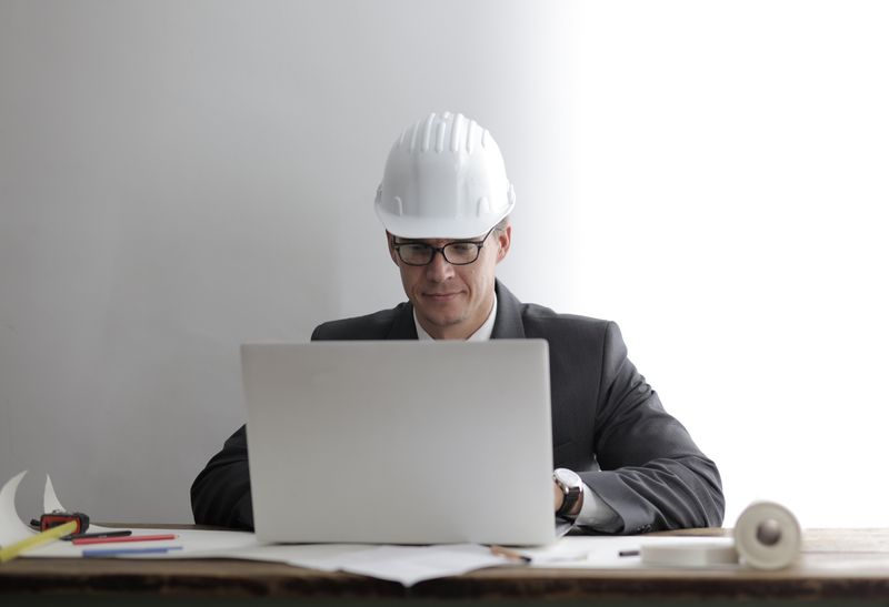 Engineer in a hard hat working on a laptop at a desk with blueprints