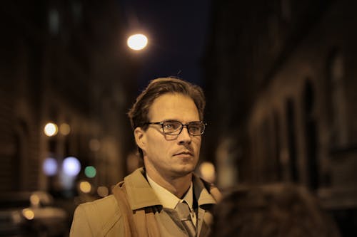 Concentrated adult male in formal clothes and eyeglasses standing on street and looking away at night