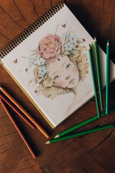 Notebook with floral illustration and colored pencils on wooden table.