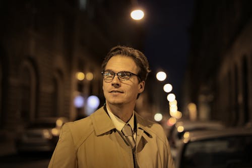 Adult positive well dressed man in eyeglasses standing on city street and looking away