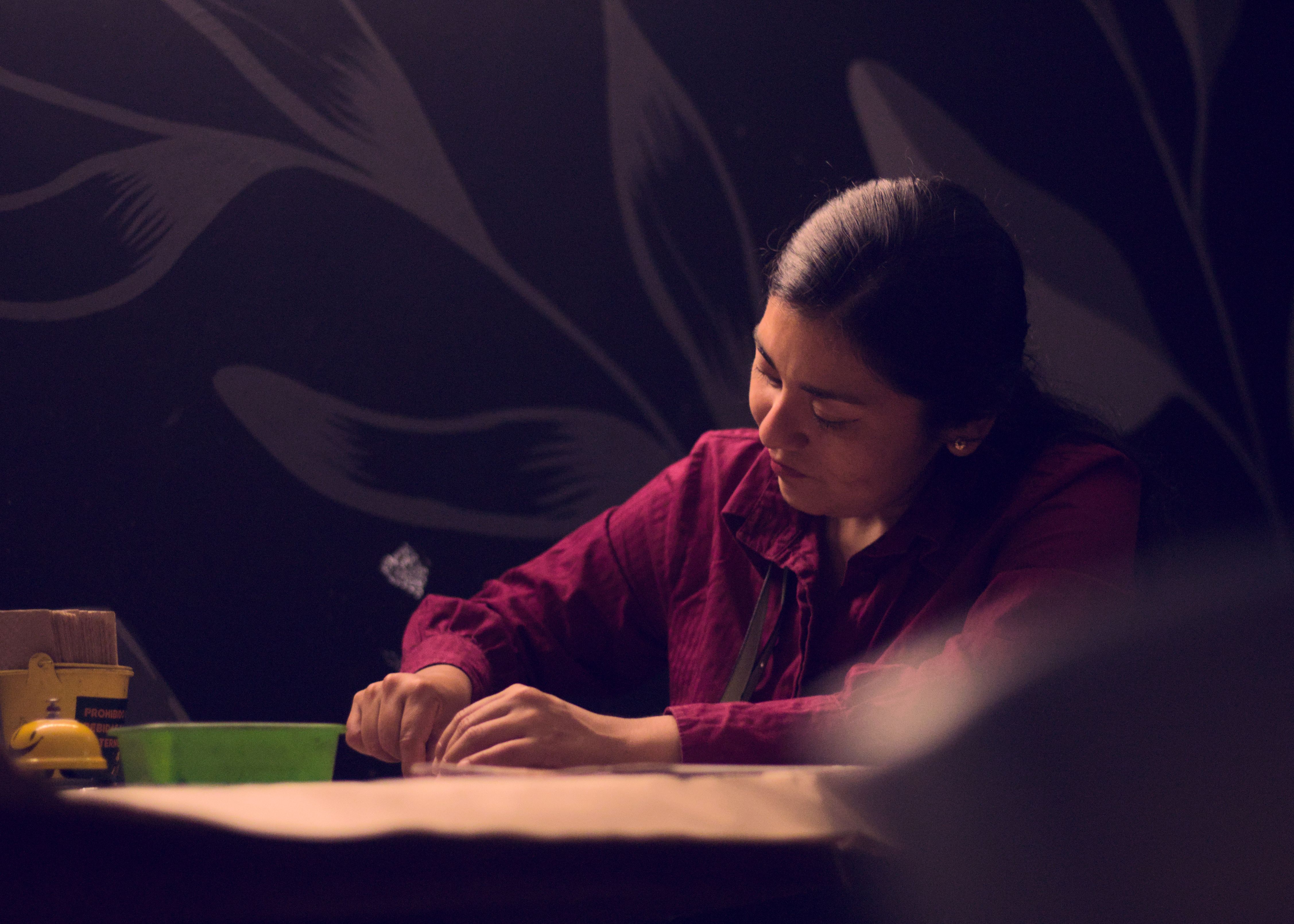Uma mulher concentra-se em desenhar em um quarto pouco iluminado, cercada por sombras melancólicas e luz suave.