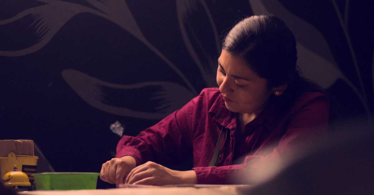 Uma mulher concentra-se em desenhar em um quarto pouco iluminado, cercada por sombras melancólicas e luz suave.
