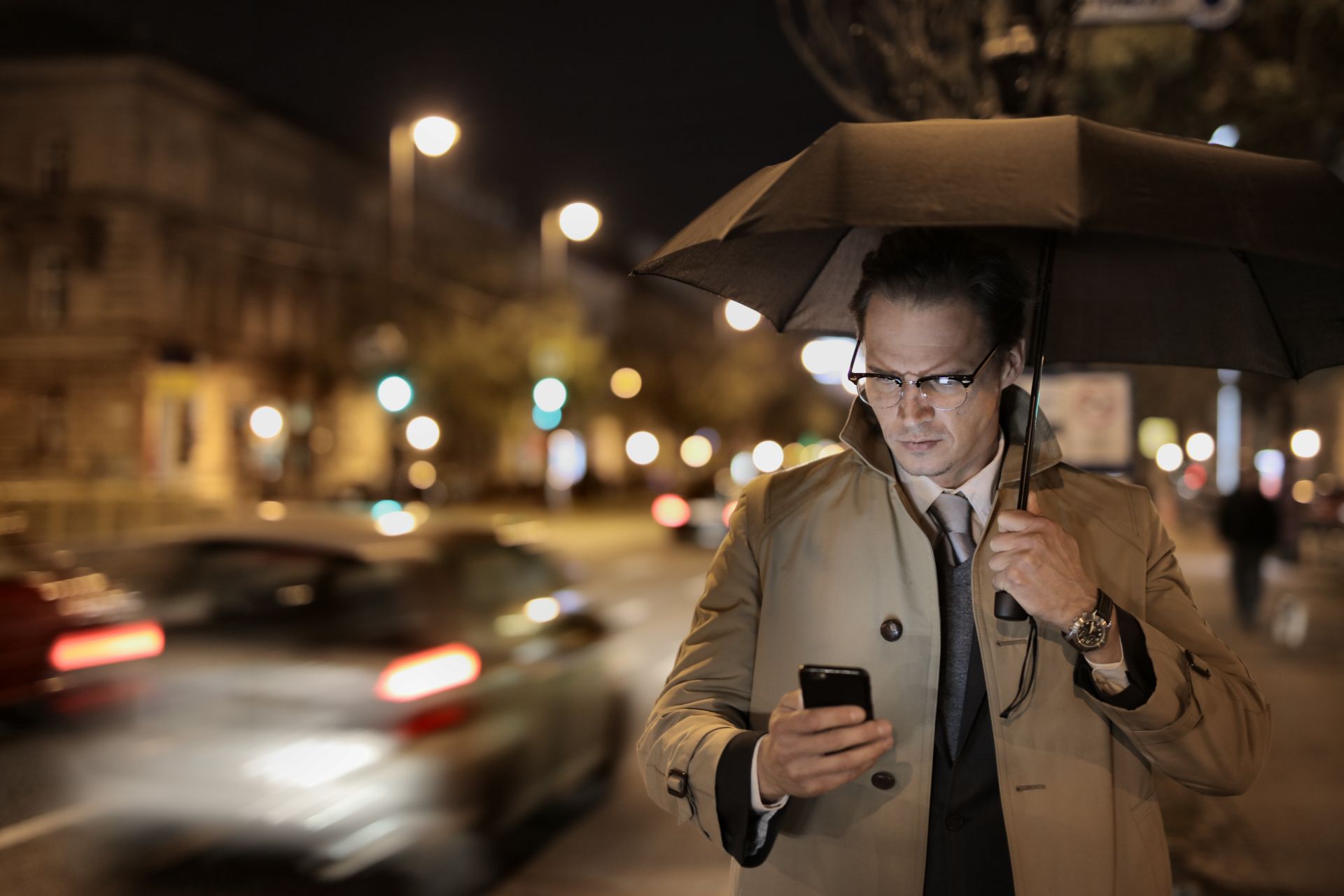 Focused formally dressed man in trench holding umbrella while standing on city street and sending message on smartphone