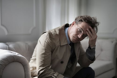 Pensive adult brown haired man in eyeglasses and trench sitting on couch and touching head while looking at camera