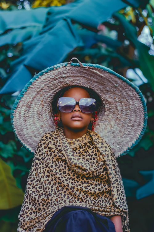Free Child in sunglasses and large straw hat poses outdoors with leopard print shawl, surrounded by lush foliage. Stock Photo