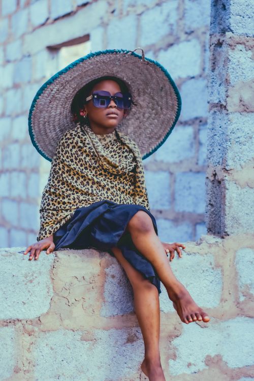 Free A young child wearing a leopard print scarf, sunglasses, and a straw hat sits on a wall in Abuja, Nigeria. Stock Photo