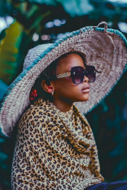 Free A child wearing a large straw hat and sunglasses, capturing a fashionable and playful moment outdoors. Stock Photo