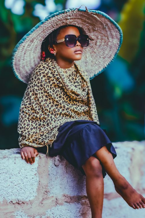 Free A fashionable child sits on a wall, wearing a leopard print shawl and wide-brimmed hat. Stock Photo