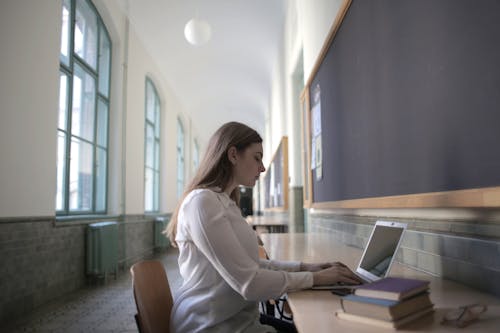 Free Side view of young brown haired concentrated woman typing on laptop while sitting at table with books Stock Photo
