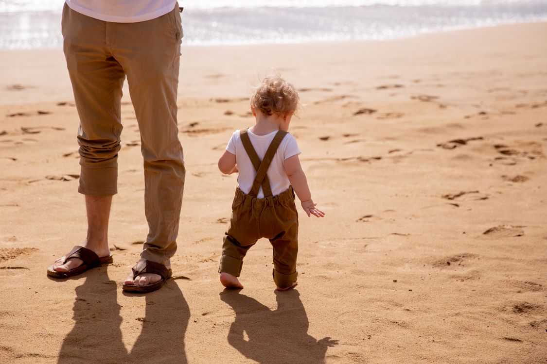 Toddler walking on sandy beach with faceless father