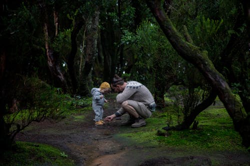 Side view of young man wearing hiking boots and warm sweater during hiking with toddler baby inside coniferous forest covered of green moss while walking on trail