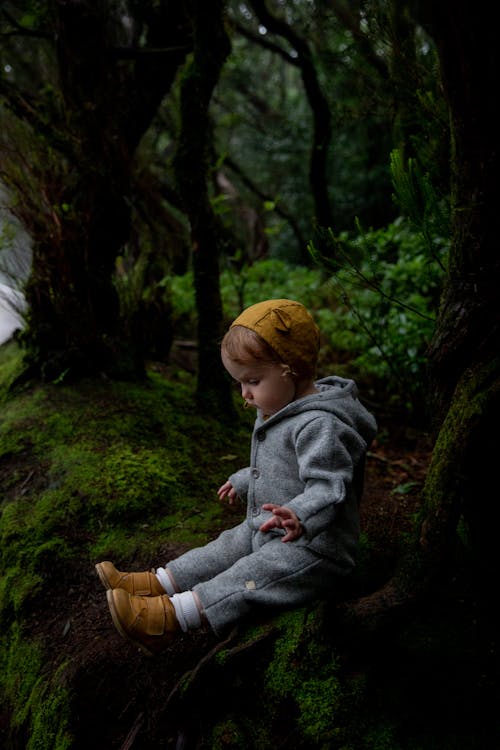 Side view of adorable toddler girl wearing warm gray bodysuit and boots sitting on ground covered with green moss in deep green forest