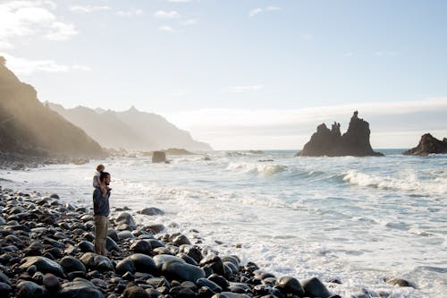 Free Side view of man carrying adorable toddler on neck while standing on pebble shore near waving sea during vacation Stock Photo